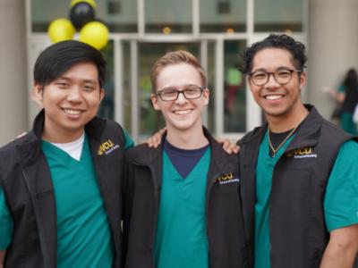 three v.c.u. nursing students wearing green scrubs and school-branded vests pose and smile for the camera at an event