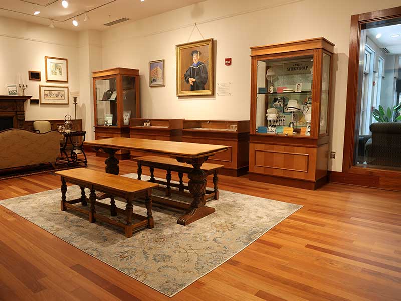 A warmly lit Heritage Room featuring a long wooden table with benches on a rug, surrounded by wood-framed display cases, portraits, and nursing memorabilia