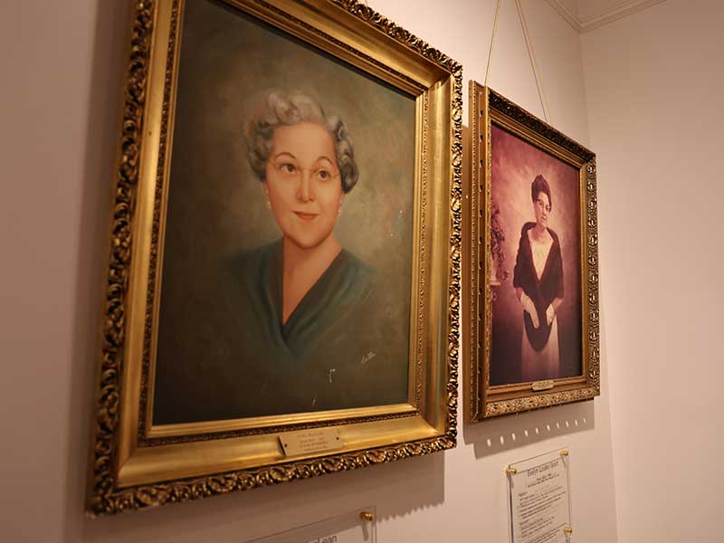 Two framed portraits of women, both in ornate gold frames. One is a painted portrait of a woman with short gray hair and a green blouse; the other is a photograph of a woman in a formal black gown.
