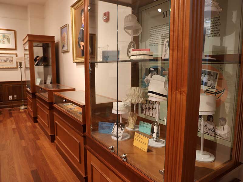 A close-up view of a wooden display case featuring historical nursing caps and educational placards, with additional portraits and artifacts visible in the background.
