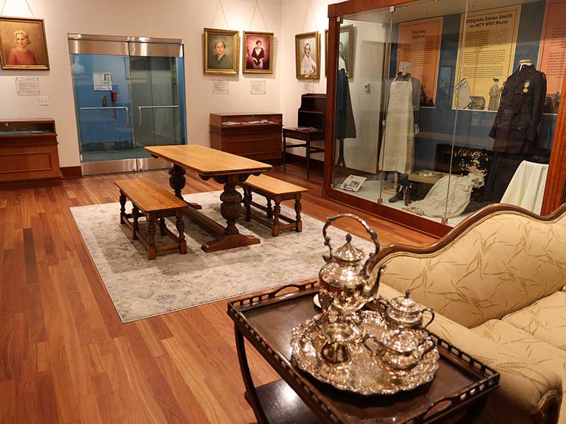 A view of the Heritage Room showcasing a silver tea set on a side table, historical nursing uniforms in a glass display case, and a central wooden table with benches beneath hanging portraits
