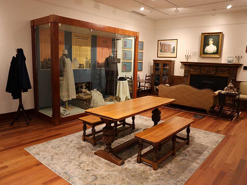A wide view of the Heritage Room showing a central wooden table with benches, a large display case with vintage nursing attire, period furniture, and framed portraits along the walls.