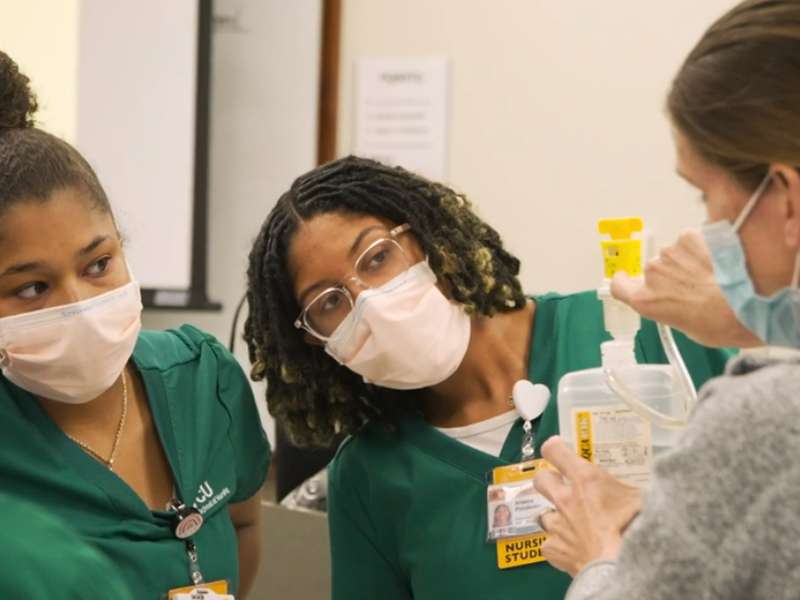two masked nursing students watch as an instructor shows them how to operate an i.v.