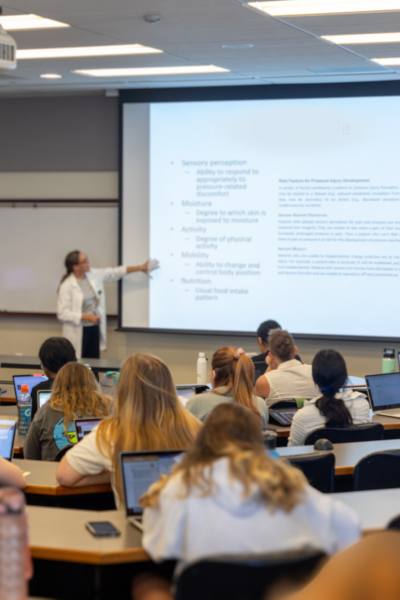 students listen and take notes on laptops in a lecture hall while a professor lectures and points to a presentation