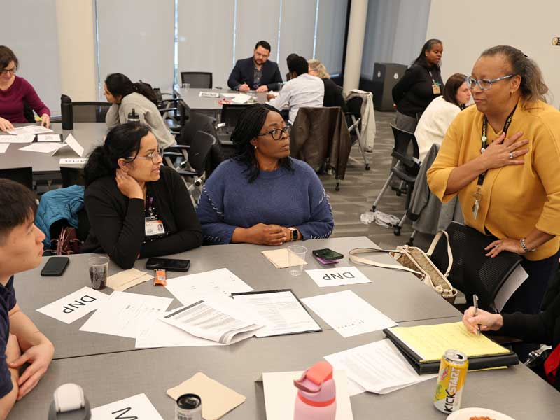A faculty member stands speaking to a group of students seated around a table with printed materials and name cards during a classroom discussion.