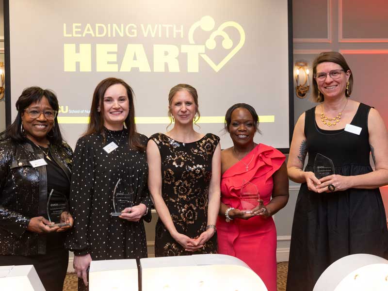 Five award recipients stand side by side holding glass awards at a V.C.U. School of Nursing ceremony, smiling in front of a screen that reads “Leading With Heart.”