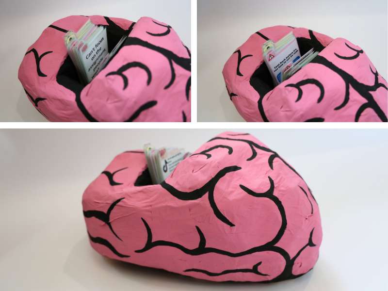 Papier-mâché sculpture of a pink brain with bold black lines mimicking folds. The top is hollowed out to hold laminated cards, some of which display mental health messages such as 'Can't focus, anxious, overwhelmed.' The piece playfully visualizes how mental load and informational overwhelm are stored in the brain.