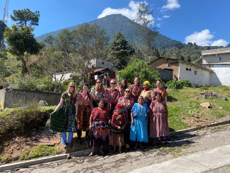 a group of seven v.c.u. nursing students, two professors, and local guatemalan women pose in traditional, colorful guatemalan garb in front of a home in a mountainous setting