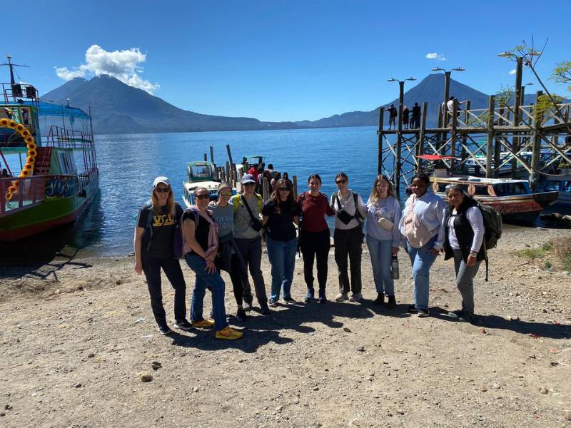 a group of seven v.c.u. nursing students with two professors and a guide posing on a beach in front of a lake with a few boats ashore