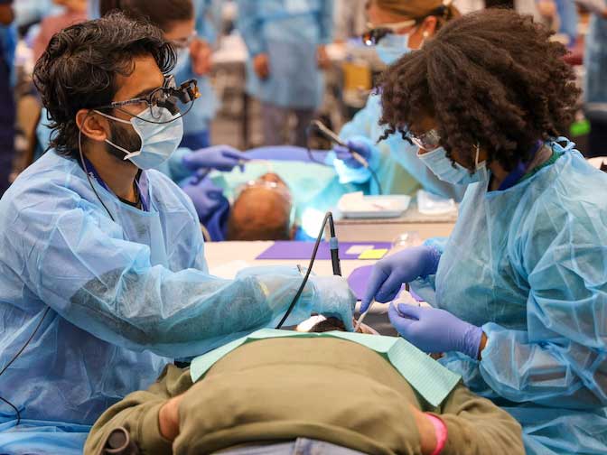 Dental students in protective gowns and masks perform procedures on patients lying on treatment tables in a large clinical training space