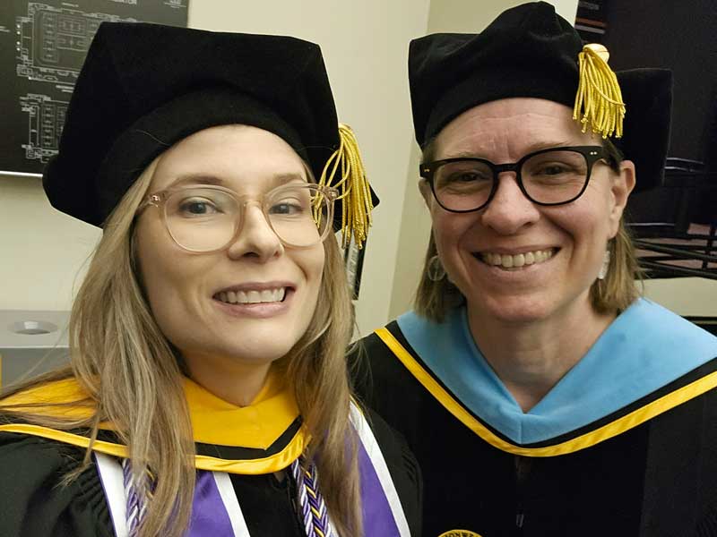 Two people wearing academic regalia smile during a graduation ceremony