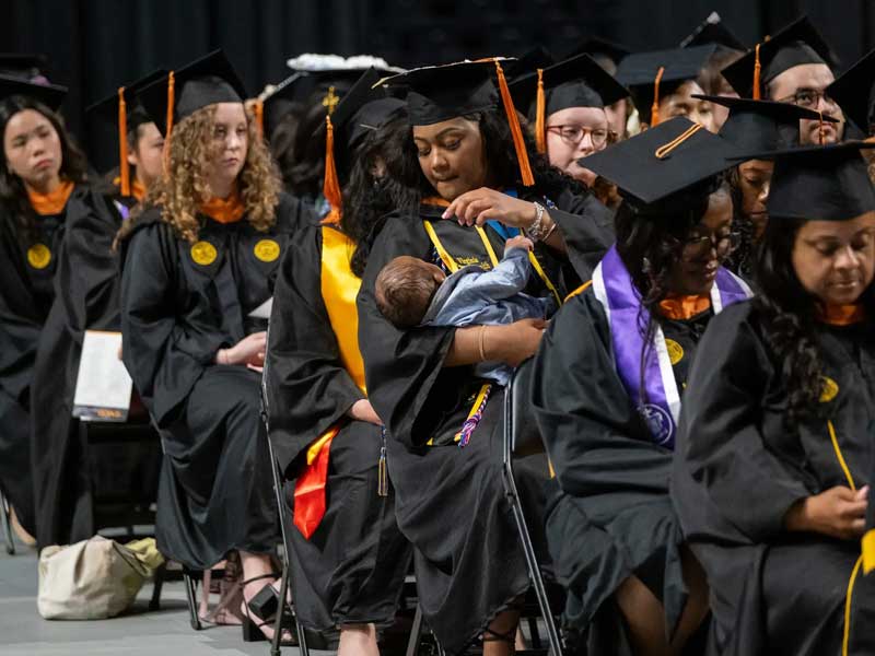a crowd of graduates dressed in v.c.u. commencement regalia sitting at a commencement ceremony - one of the graduates holds her infant son