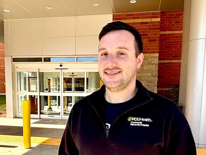 Evan Hundley wearing a black V.C.U. Health Community Memorial Hospital jacket stands outside the hospital entrance, smiling at the camera