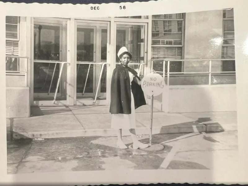 charlotte pollard in a photo from 1958 where she is dressed in a nurse uniform in front of a no parking sign in a parking lot