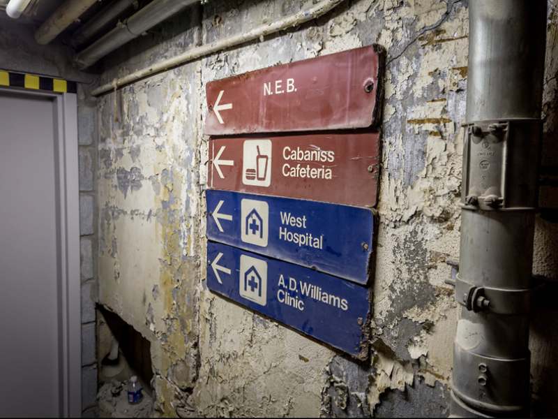 a dimly lit underground tunnel with peeling paint on the walls and directional signage reading 'n.e.b., cabaniss cafeteria, west hospital and a.d.williams clinic'