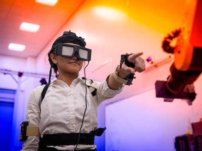 A student wearing virtual-reality goggles and motion-capture gear reaches toward a robotic arm in a brightly lit lab