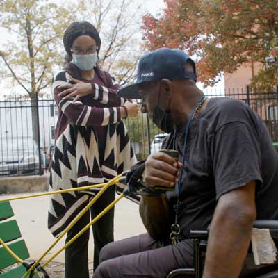 a person in a wheelchair performs arm strengthening exercises with resistance bands attached to an outdoor bench at the behest of a community health worker giving guidance