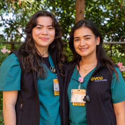 Two nursing students wearing green scrubs and black vests with VCU School of Nursing logos smile while standing outdoors. They both have ID badges and stethoscopes, with trees and flowers in the background.