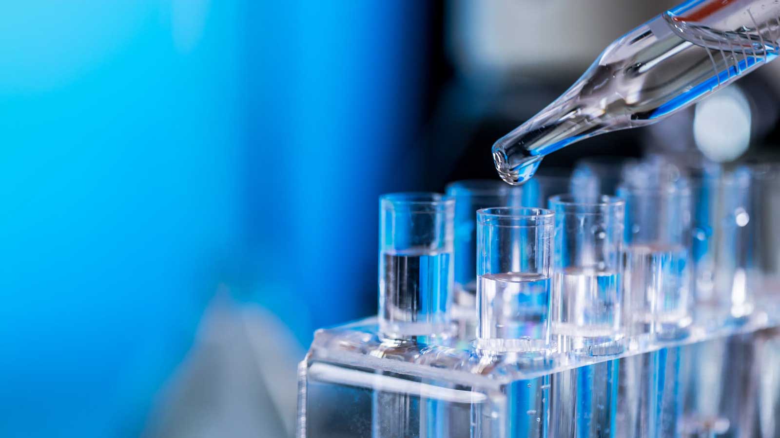 A pipette dispenses a drop of clear liquid above test tubes containing clear liquid in a laboratory rack