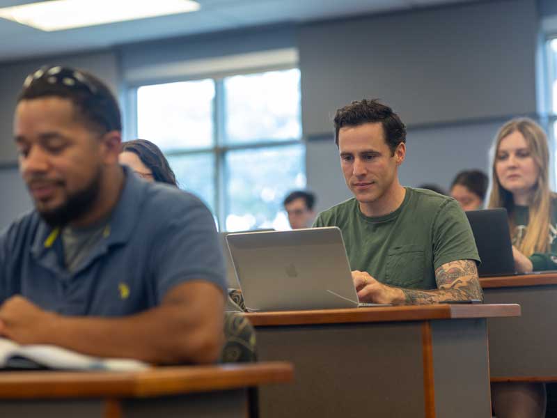 students seated in a lecture hall taking notes on laptop computers