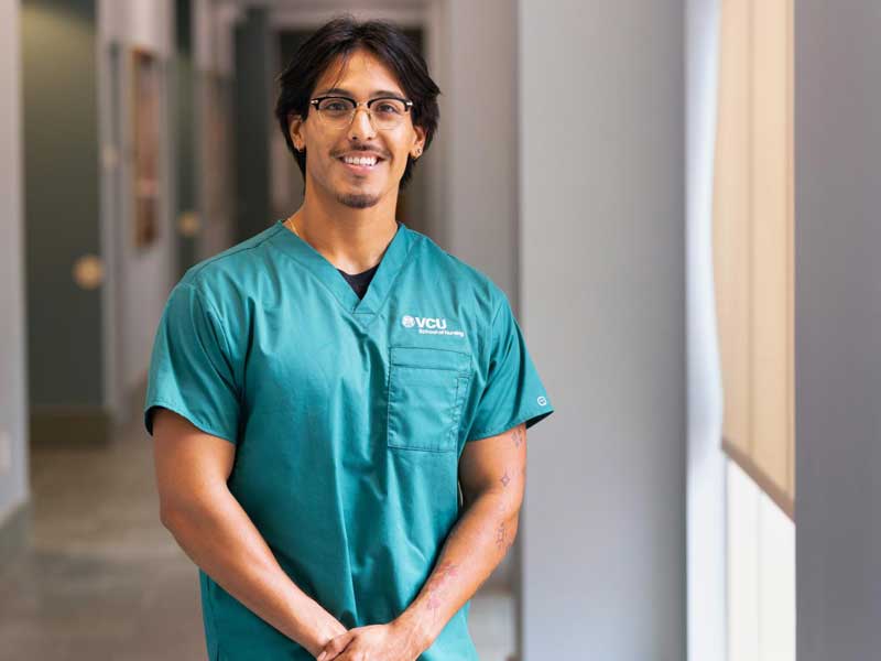 A student wearing green V.C.U. School of Nursing scrubs stands in a hallway with hands folded, smiling toward the camera