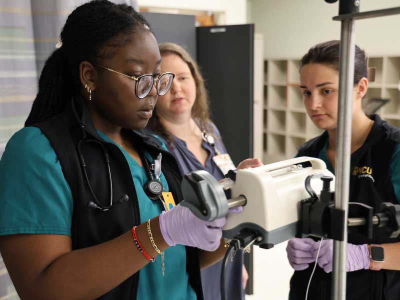 A nursing student practices using clinical equipment under instructor supervision during a simulation lab.