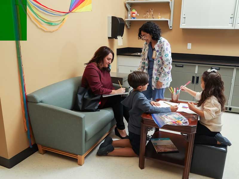 a mother signs a form in a clinic waiting room while a nurse looks on and her two children play at a nearby art table