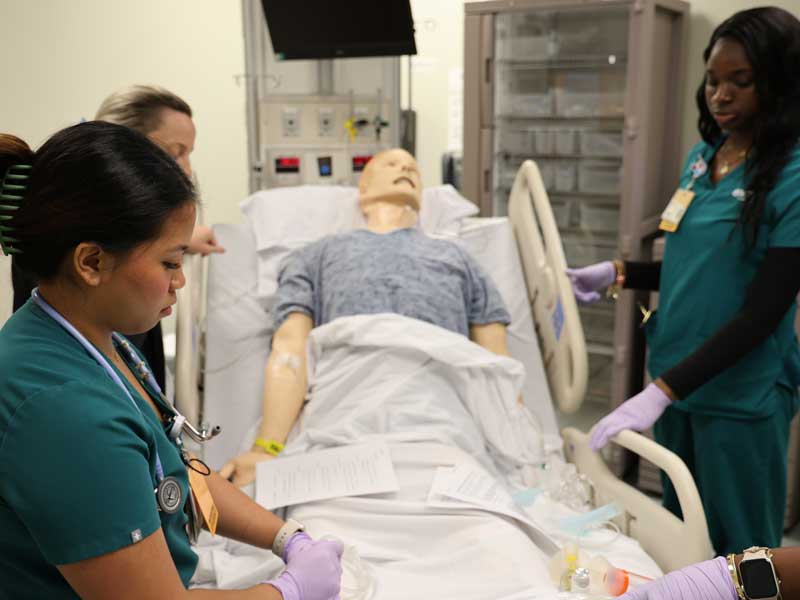 Nursing students work with an adult patient simulation mannequin in a hospital bed during clinical training